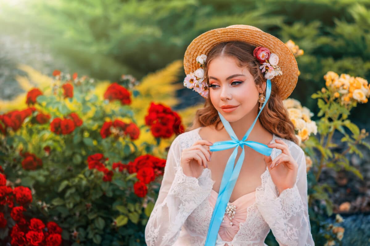 Fantasy girl princess beautiful face, delicate make-up, sttraw hat on head, long hair blue ribbon. Happy woman sits in garden enjoys nature green bushes red flowers roses. art pink white vintage dress