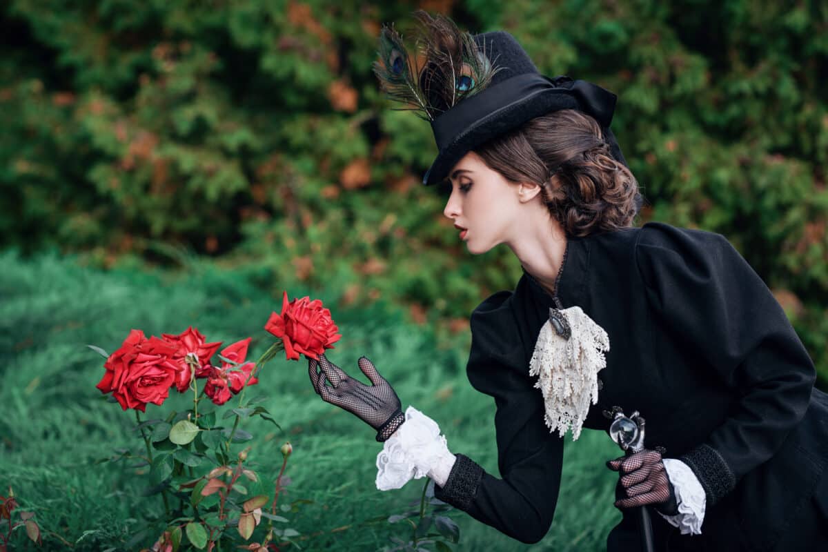 a young aristocrat lady in black vintage clothes touches the red roses in the garden