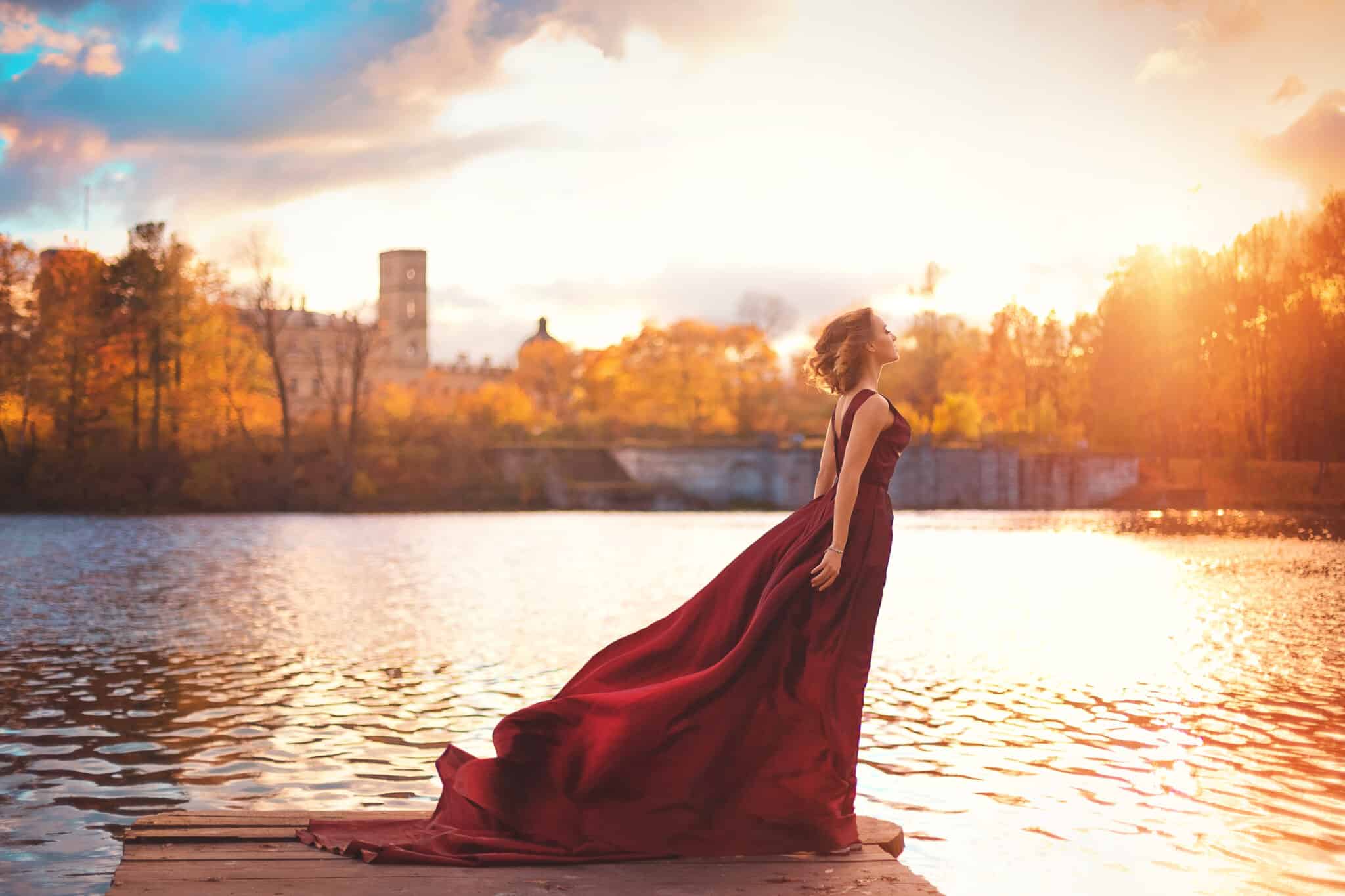 Romantic young woman in a long red dress standing by the river
