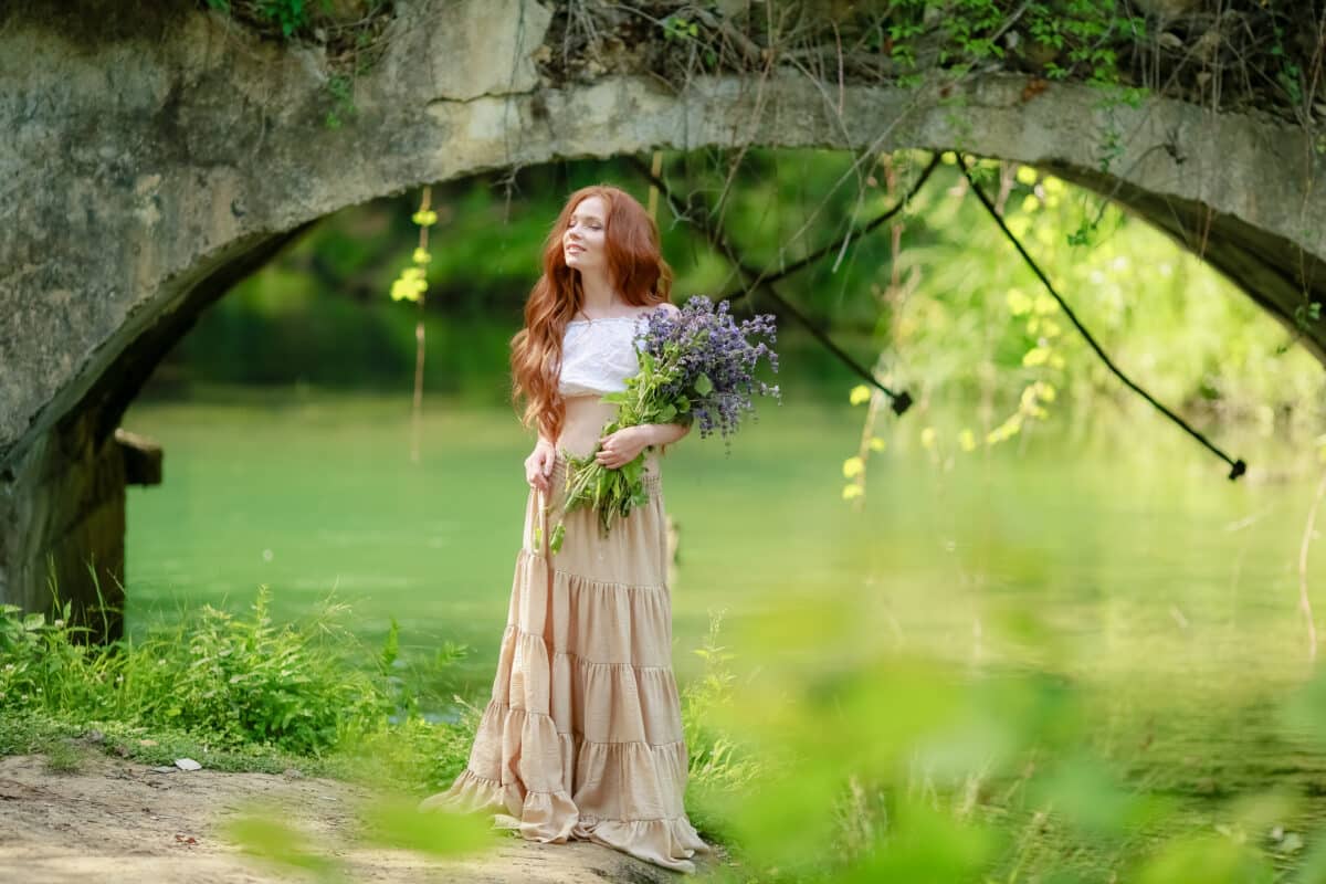 A woman in a long dress walks near the forest old bridge over the river