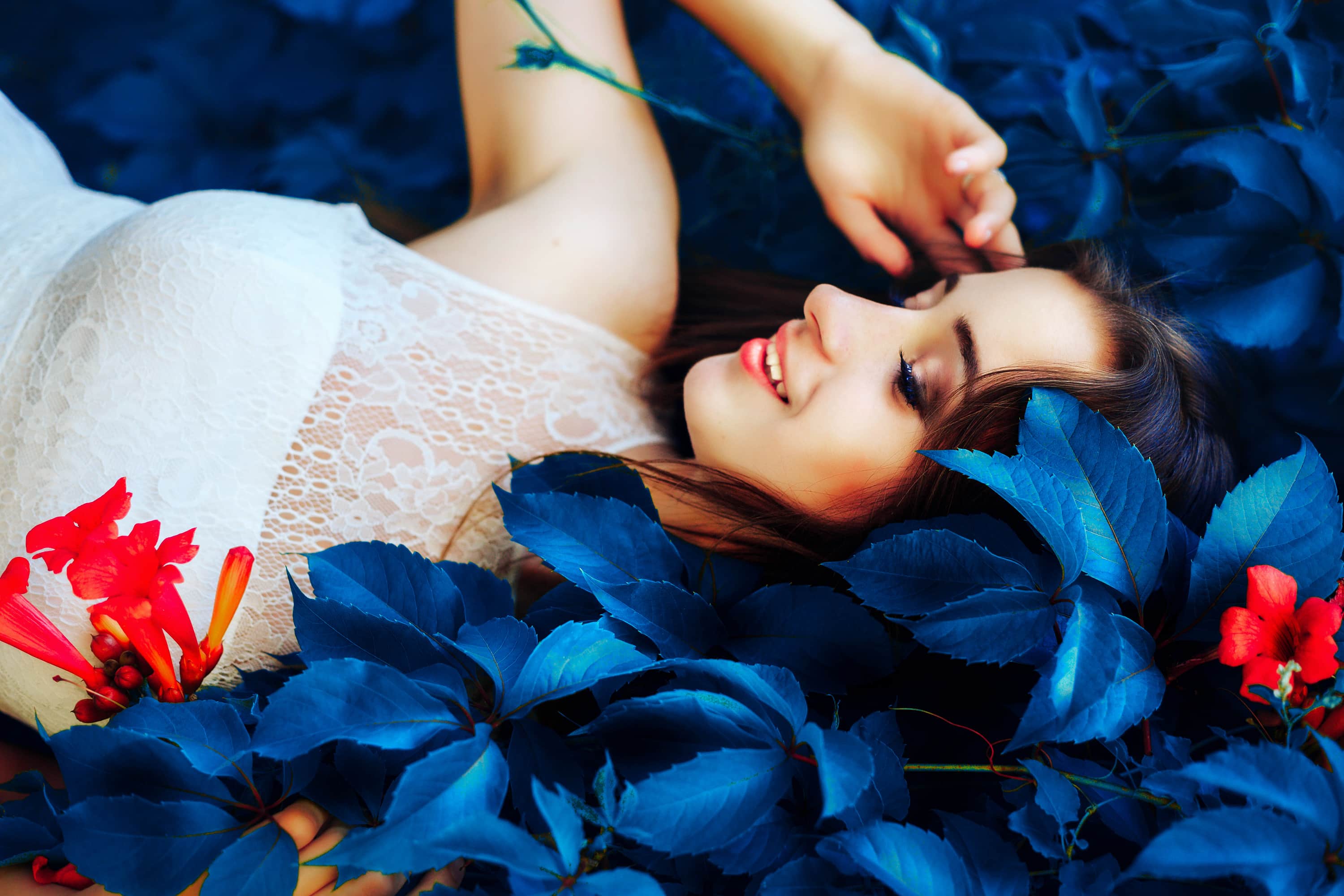 Beautiful young brunette woman in white dress lies on fantastic blue color leaves and flowers