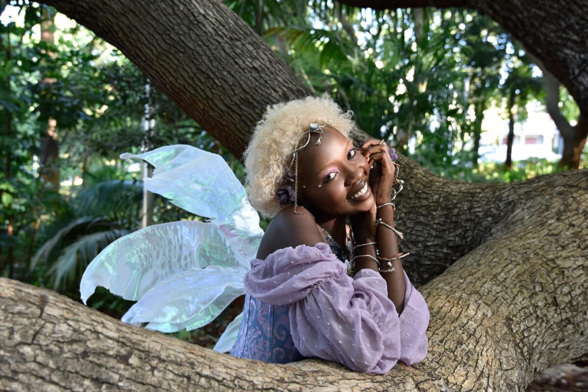 Portrait of beautiful African woman wearing purple fantasy costume, magical fairy wings & flower crown afro, climbing tree branches in forest location with natural glowing lighting.