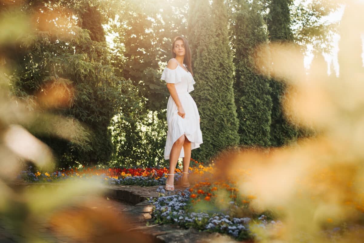 a young woman with dark skin in white light summer dress standing by flower bed at sunset