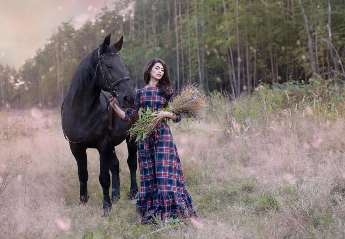 Art photo: romantic girl in a Scottish dress and a bouquet of flowers with a black stallion