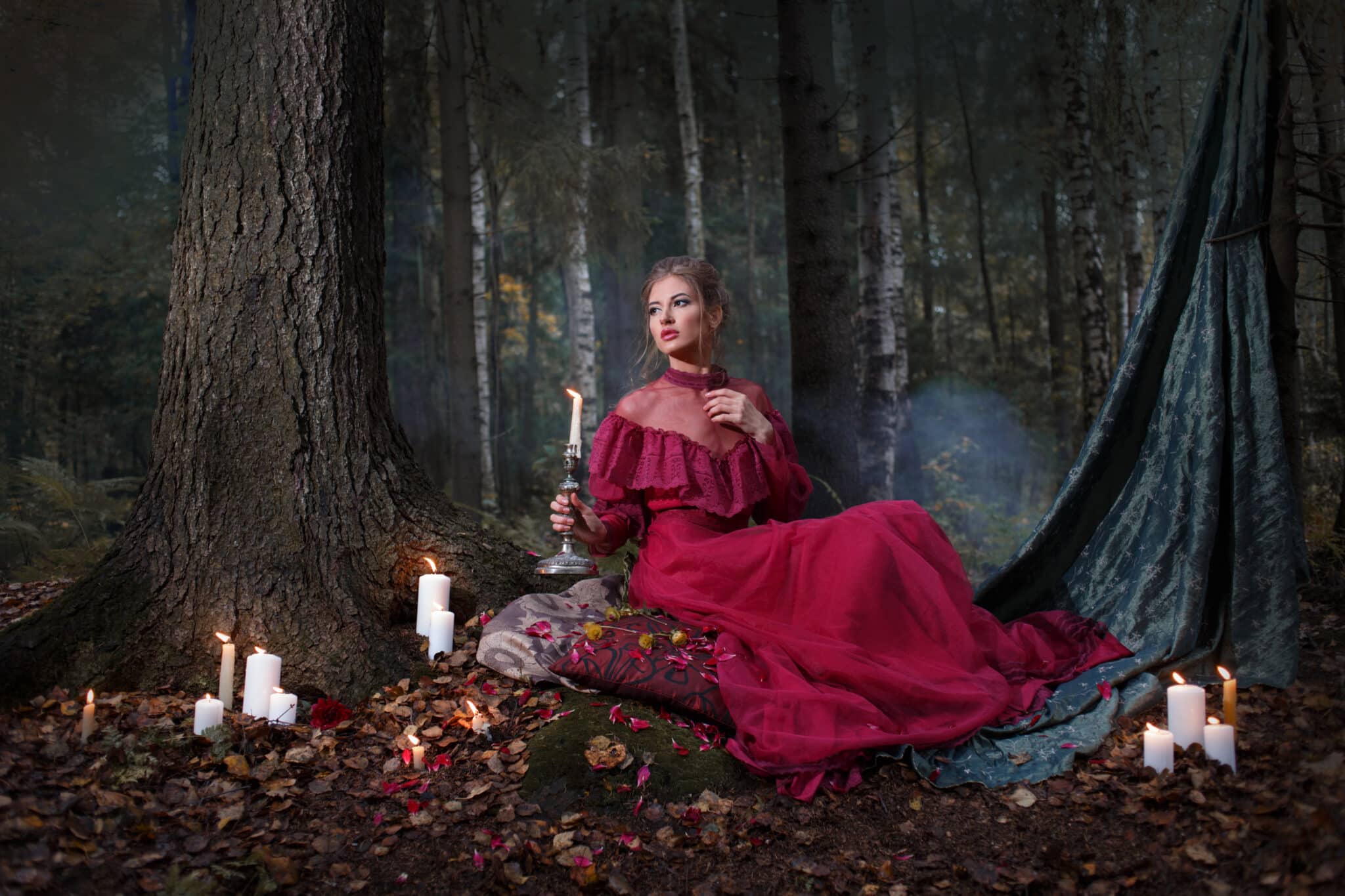 a beautiful woman in a burgundy vintage dress holding a candle sitting in a mysterious forest with lit candles beside her