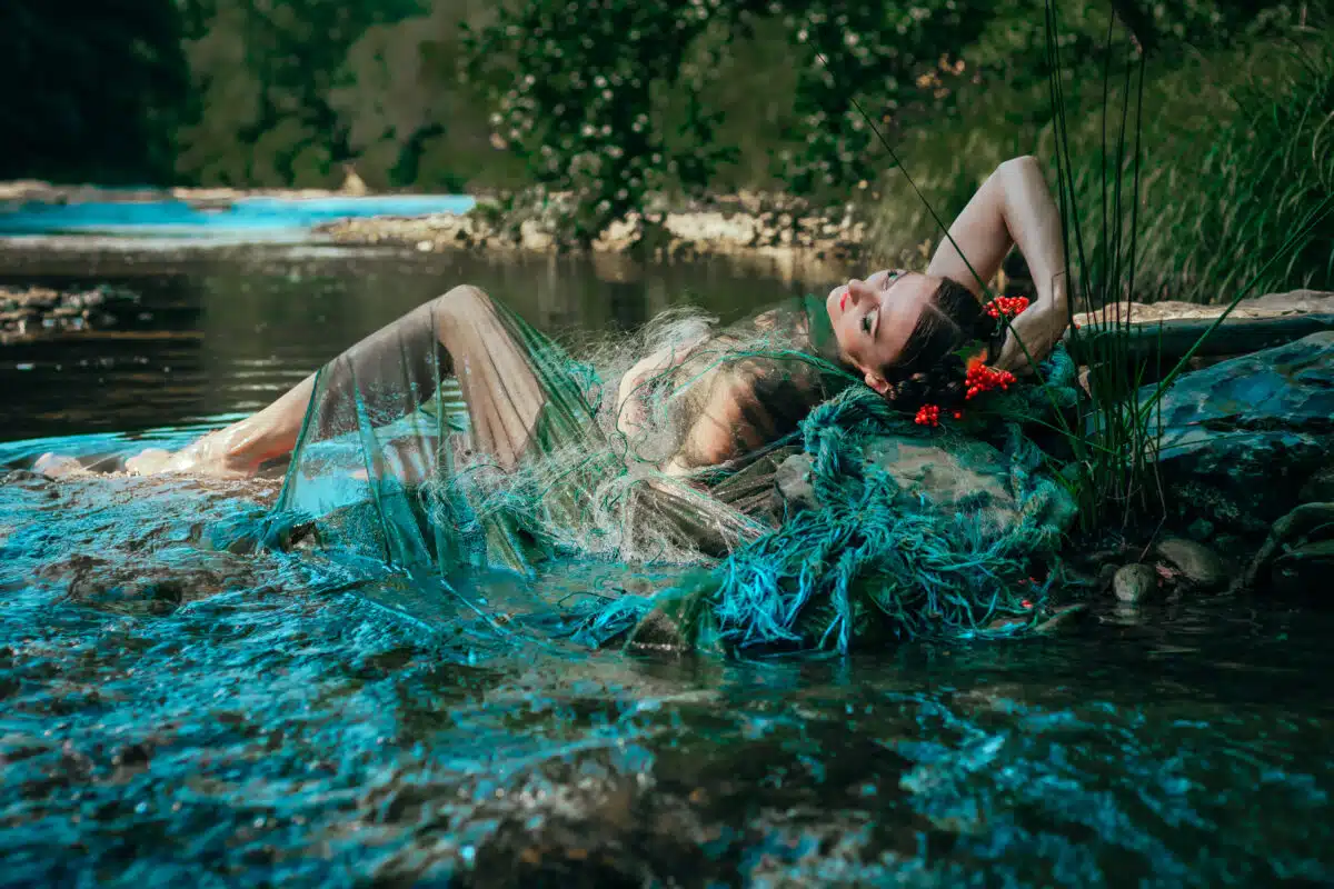 A beautiful young woman with a fabulous semi-transparent dress of a mermaid or nymph poses on a summer day on the rocks by a forest river.