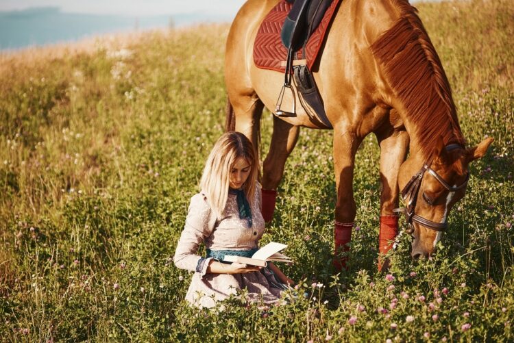 girl sitting on the grass reading a book in the field next to a horse