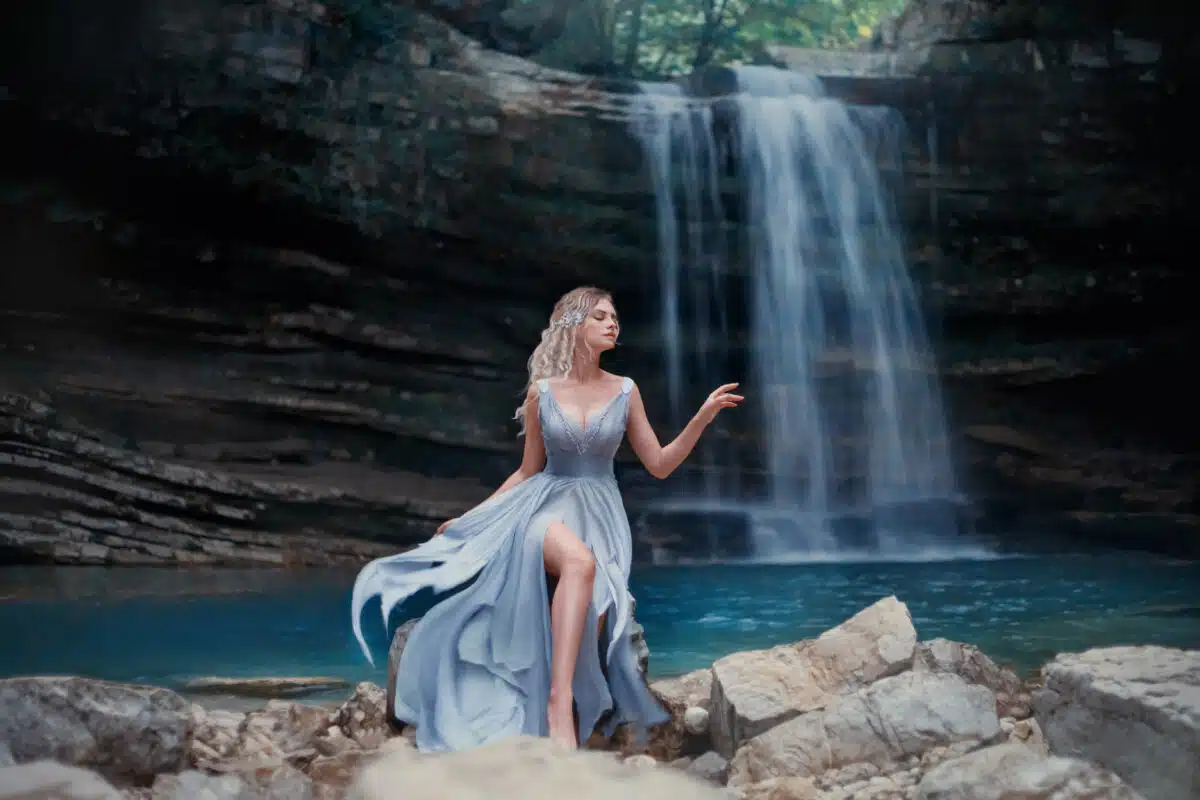 A curly blonde girl in a luxurious blue dress sits on white stones against the backdrop of a fabulous landscape. River Mermaid near the lake with a waterfall.
