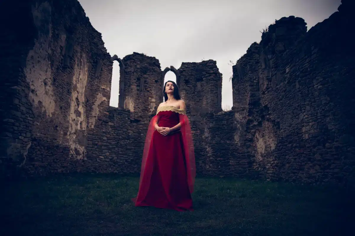 Caucasian woman wearing a medieval red dress posing against historical ancient fortifications