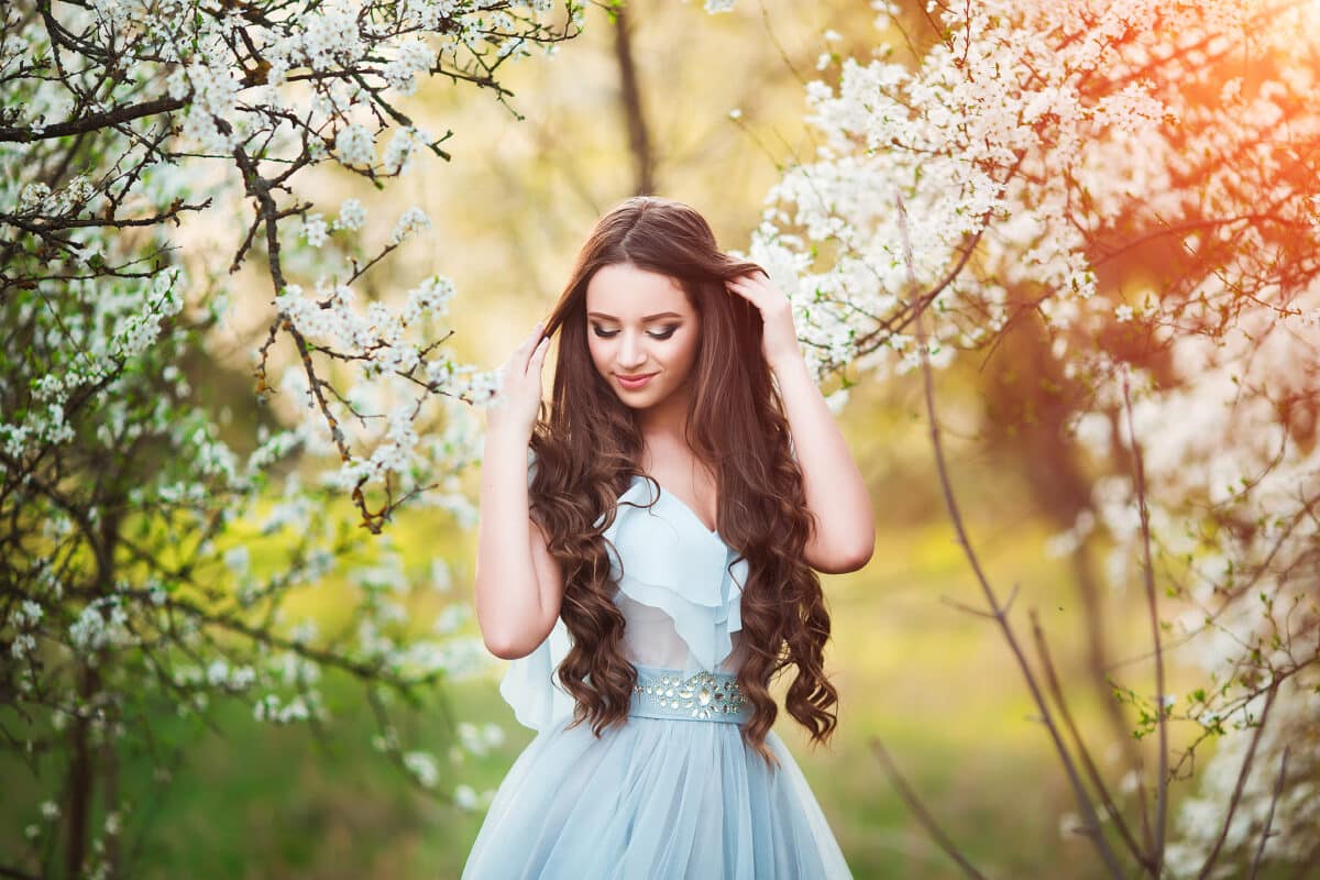 Happy beautiful young woman with long black healthy hair enjoy fresh flowers and sun light in blossom park at sunset.