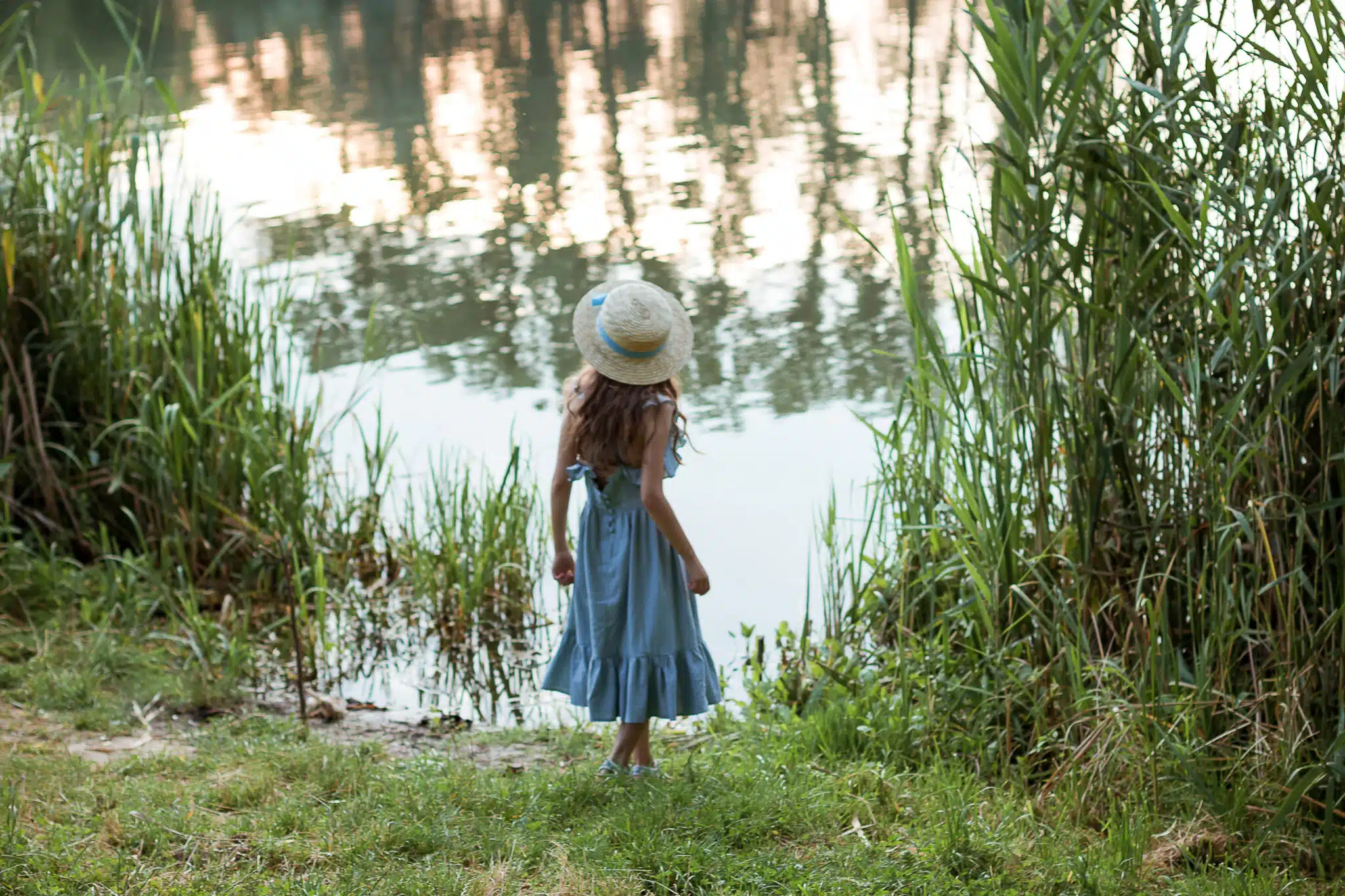 A girl in blue dress and straw hat stands on the shore of the lake