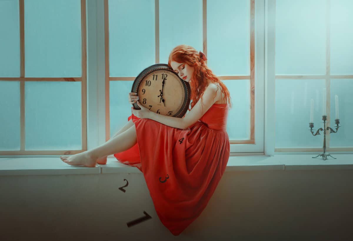 Redhead fantasy princess in a red dress is sitting on the windowsill hugging a huge clock