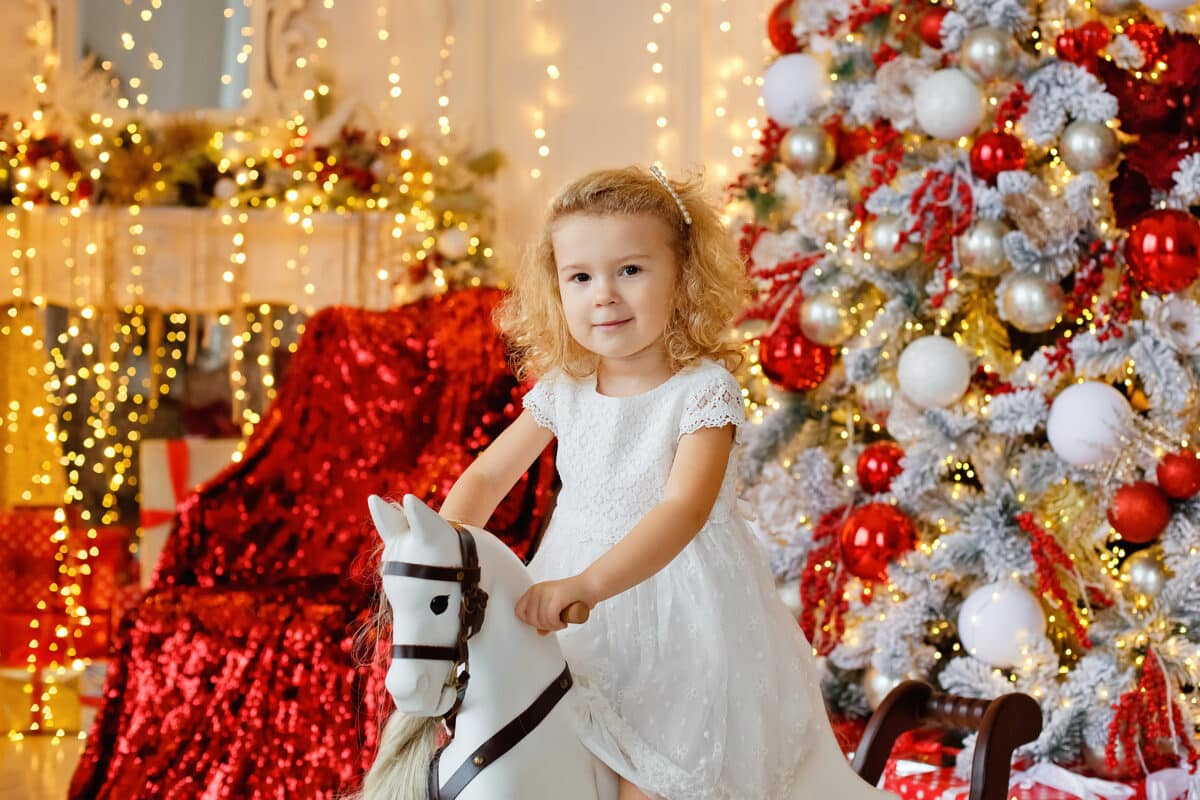 A small smiling curly-haired blonde girl in a white dress is sitting on a rocking horse 