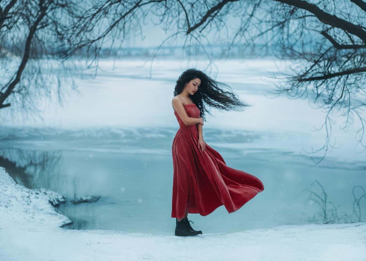 sad young brunette woman in a red dress standing by the frozen lake in winter forest