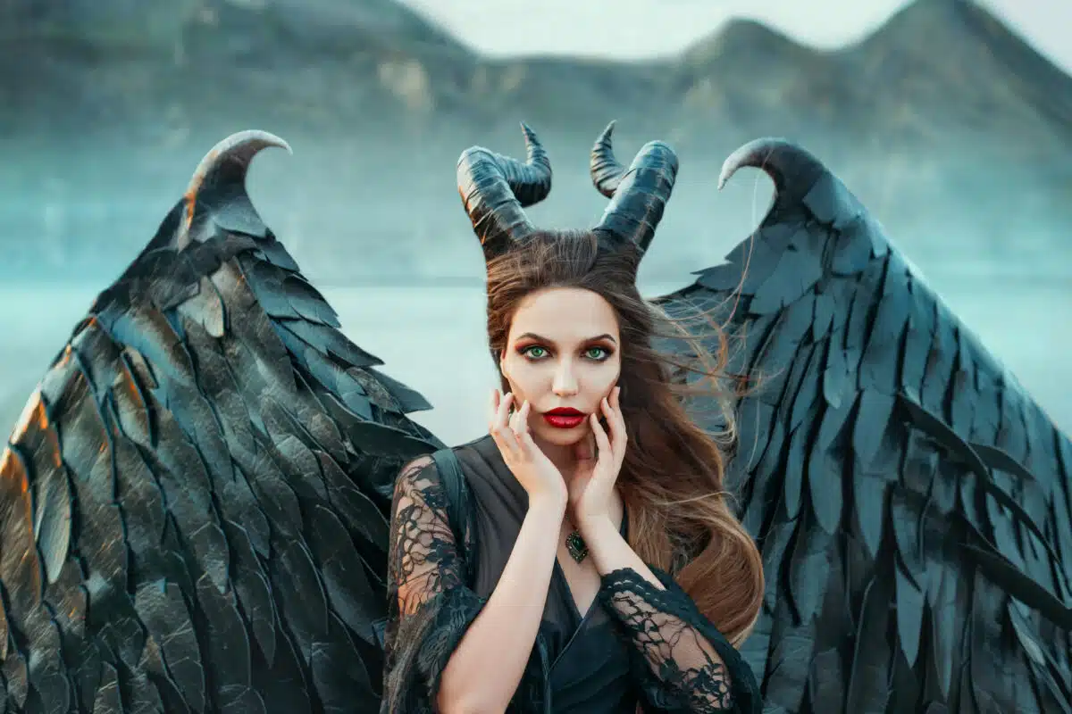 charming portrait of dark angel with sharp horns and claws on strong powerful wings, wicked witch in black lace dress brought hands to face, bright red lipstick and green eyes, art photo in blue shade