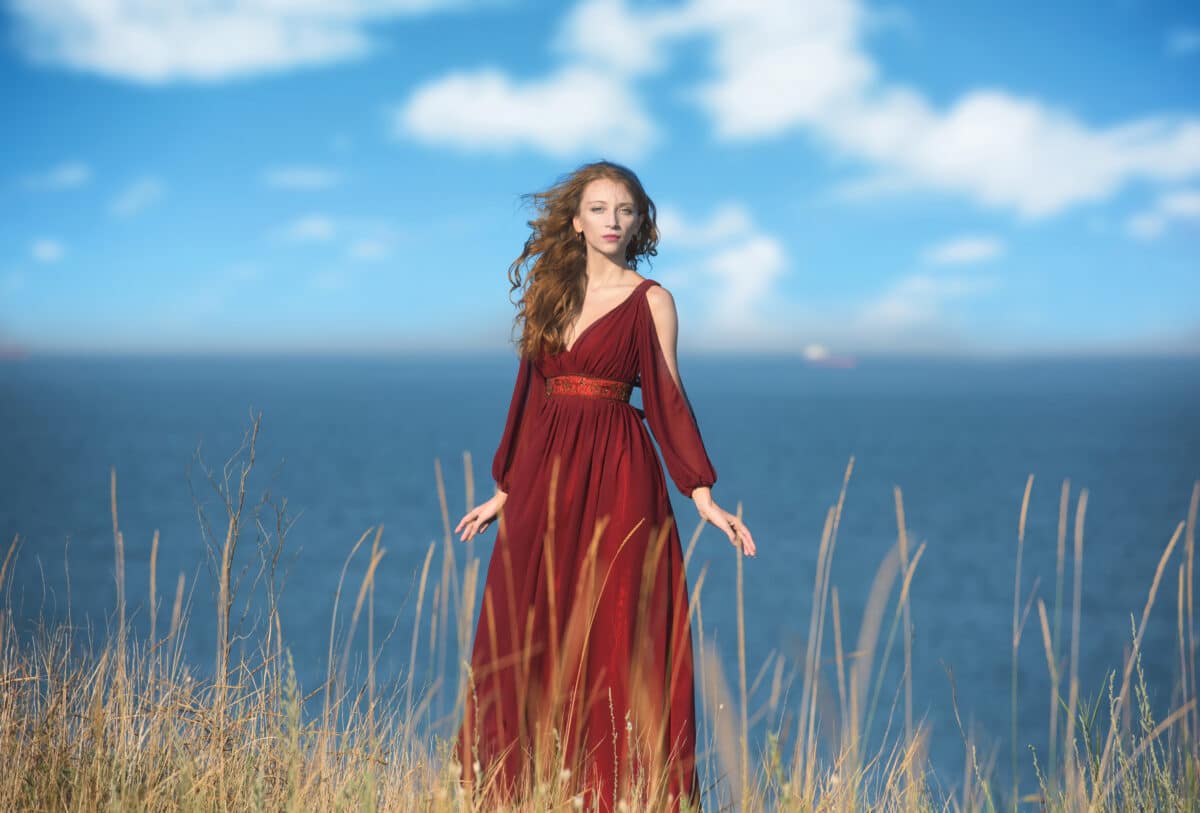 enchanting maiden in red flowy dress dress standing on a mountain by the ocean
