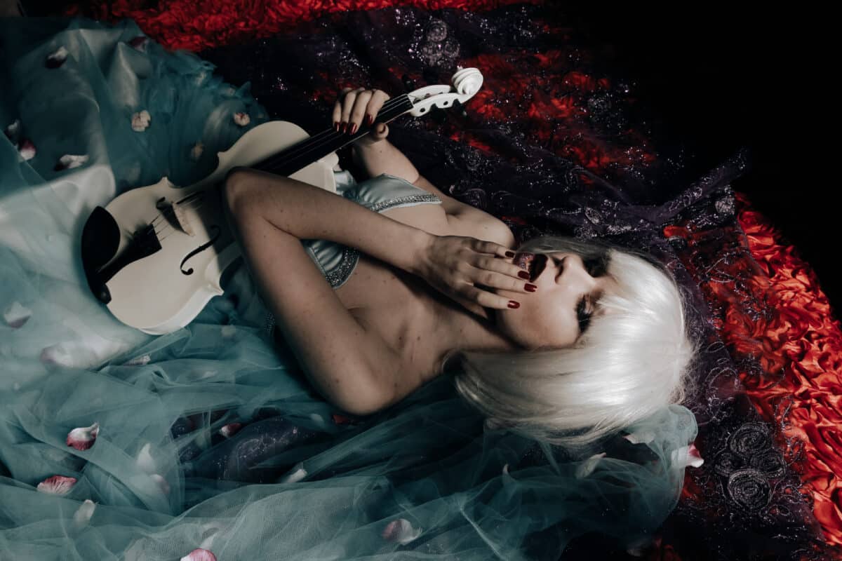 young blonde woman with dark make up holding a white violin lying on the bed with wilted petals
