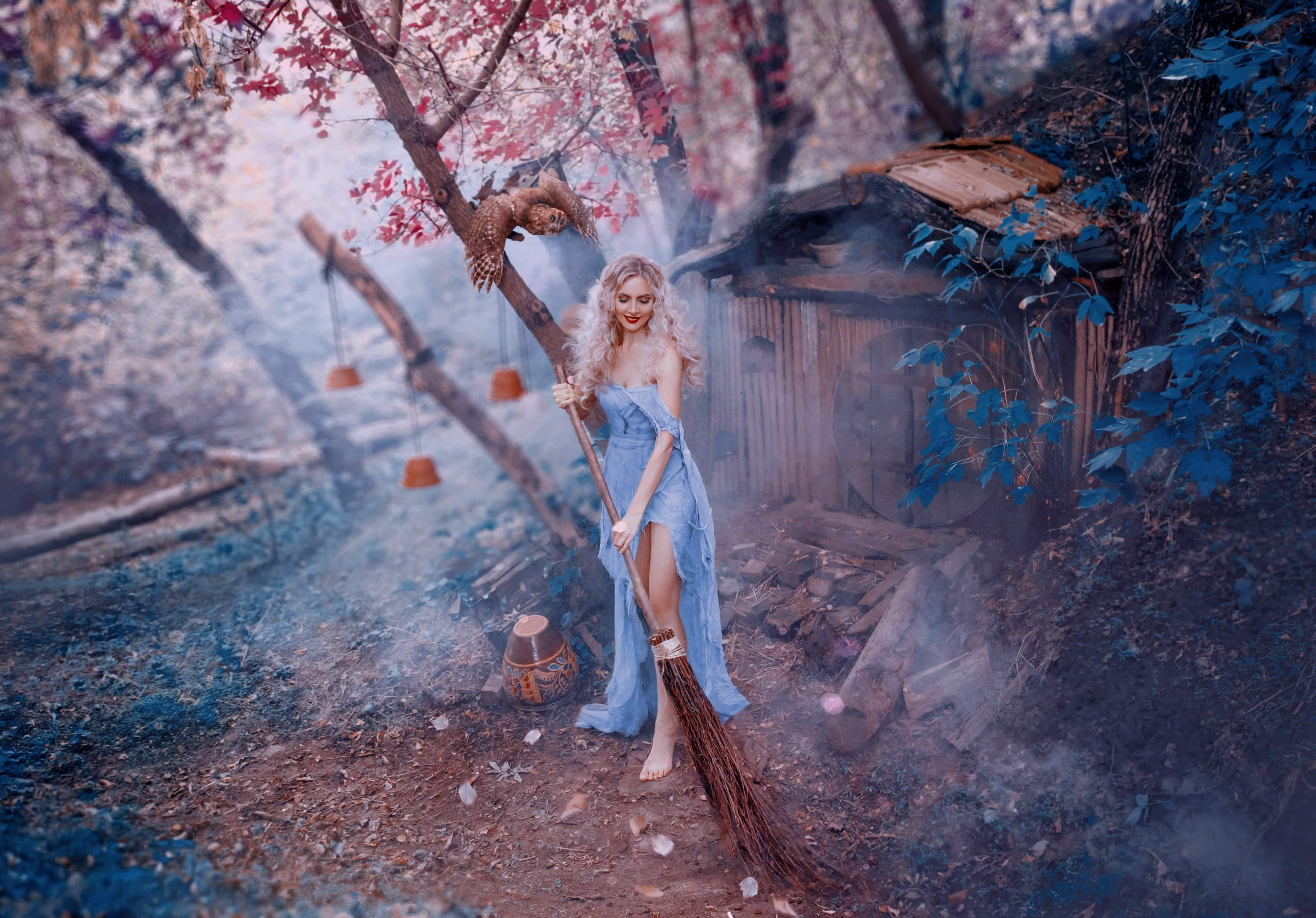 attractive blond lady in long light dress of thin fabric with naked shoulder and open legs sweeps leaves with broom near her little forest wooden house. charming kind witch resting from the magic
