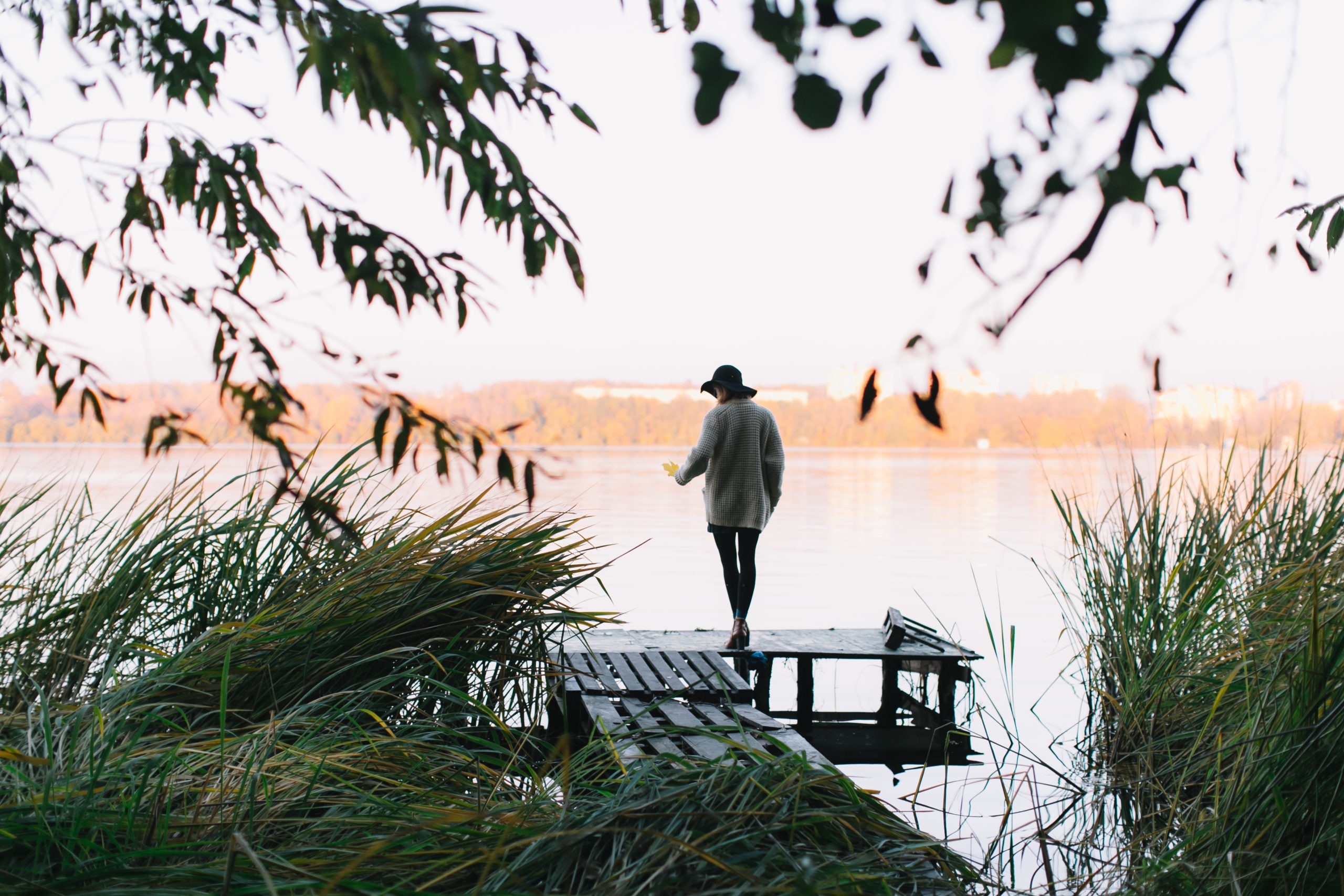 Melancholic woman standing by the lake on a wooden dock, tall grasses in the background.