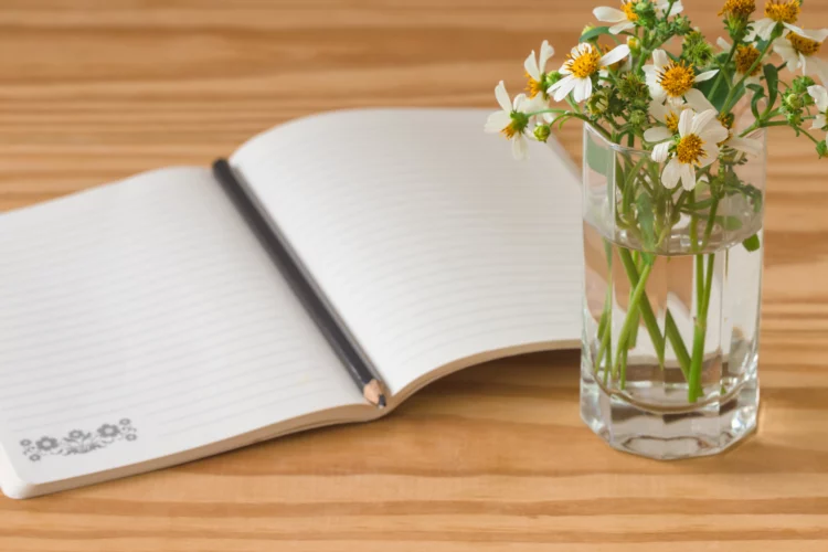 White notebook on rustic wood table with black pen and a glass with flowers.