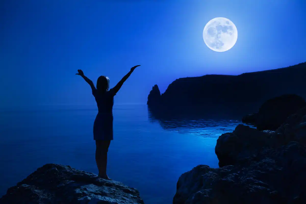 mysterious woman standing by the blue ocean with the majestic moon in the distance