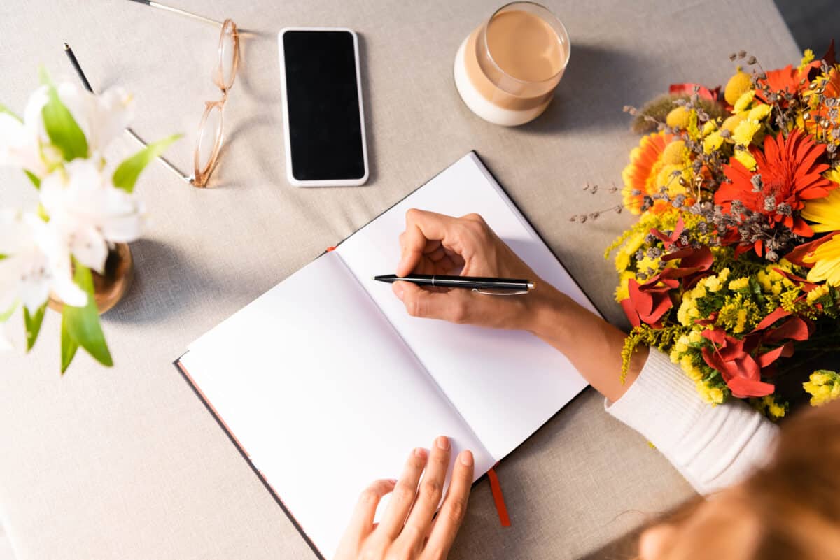 cropped view of woman writing in notepad in cafe with coffee, smartphone and flowers