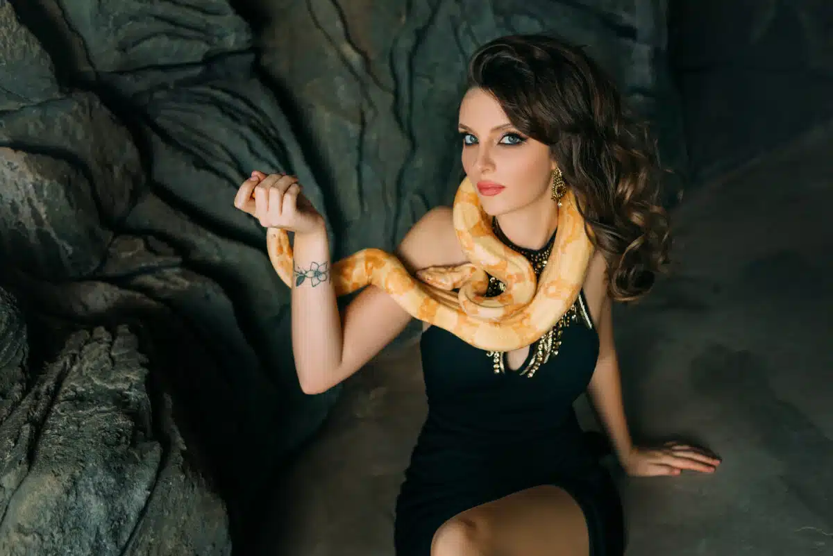 beautiful woman sits on gray stone natural cave holding a yellow snake coiling around her neck