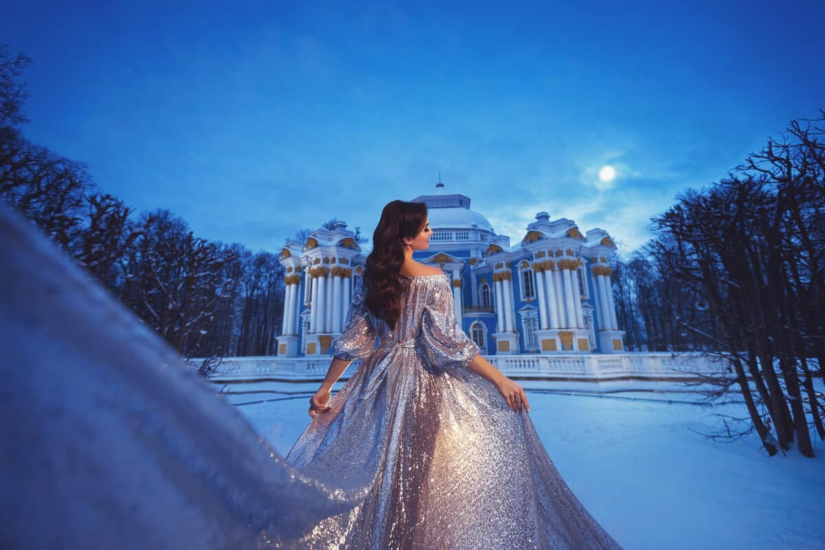 A beautiful young girl in a shiny silver dress with a long train on the background of a snow-covered palace park. A magical night portrait.