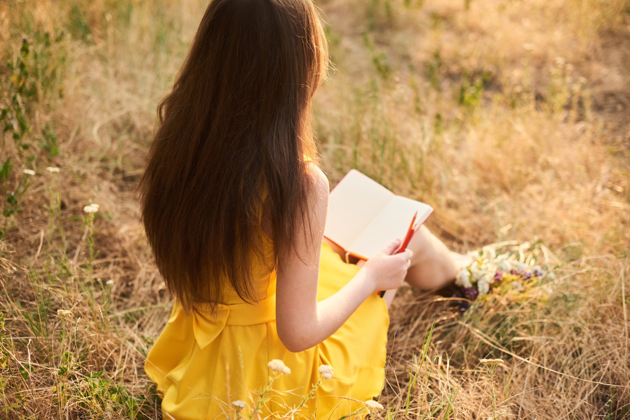 Melancholic long-haired blond girl sits with her back on the lawn with a notebook and pen.