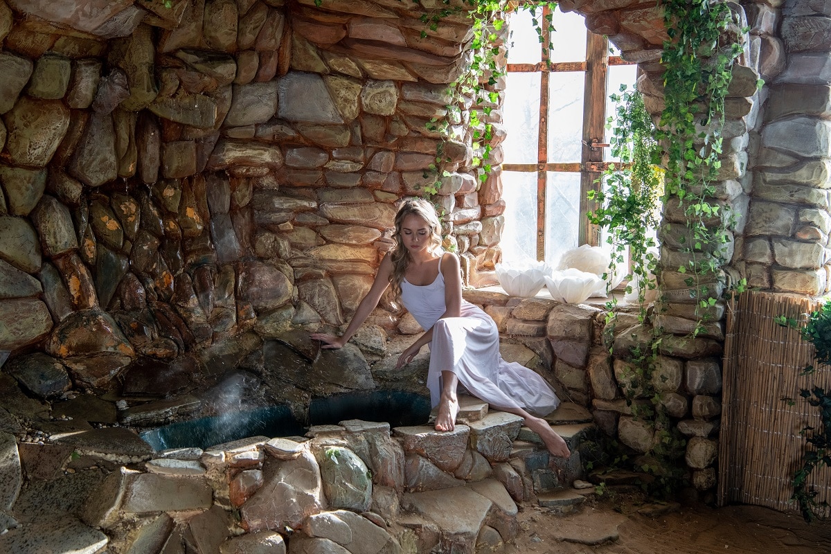 beautiful blonde in a white dress sits near a stone wall by the well