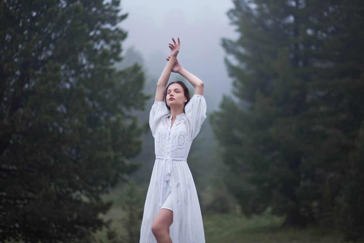 beautiful lady in a white dress is standing in the misty forest with arms raised heavenwards
