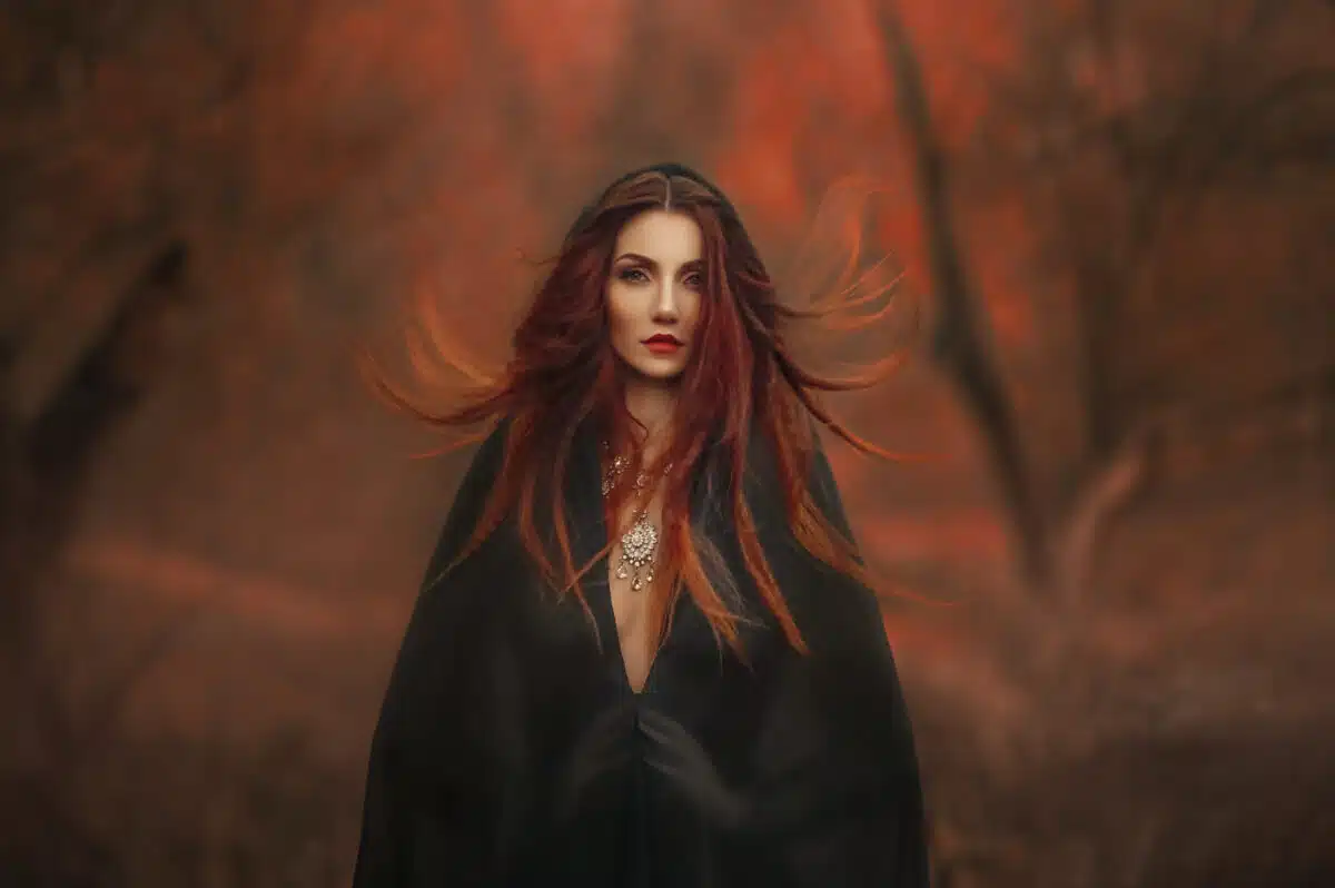 fantasy gothic woman dark witch. Red-haired evil Girl demon in black dress cape hood. Long hair flutters fly in wind. Dark dense deep autumn forest orange colors trees. Medieval dress, silk clothes