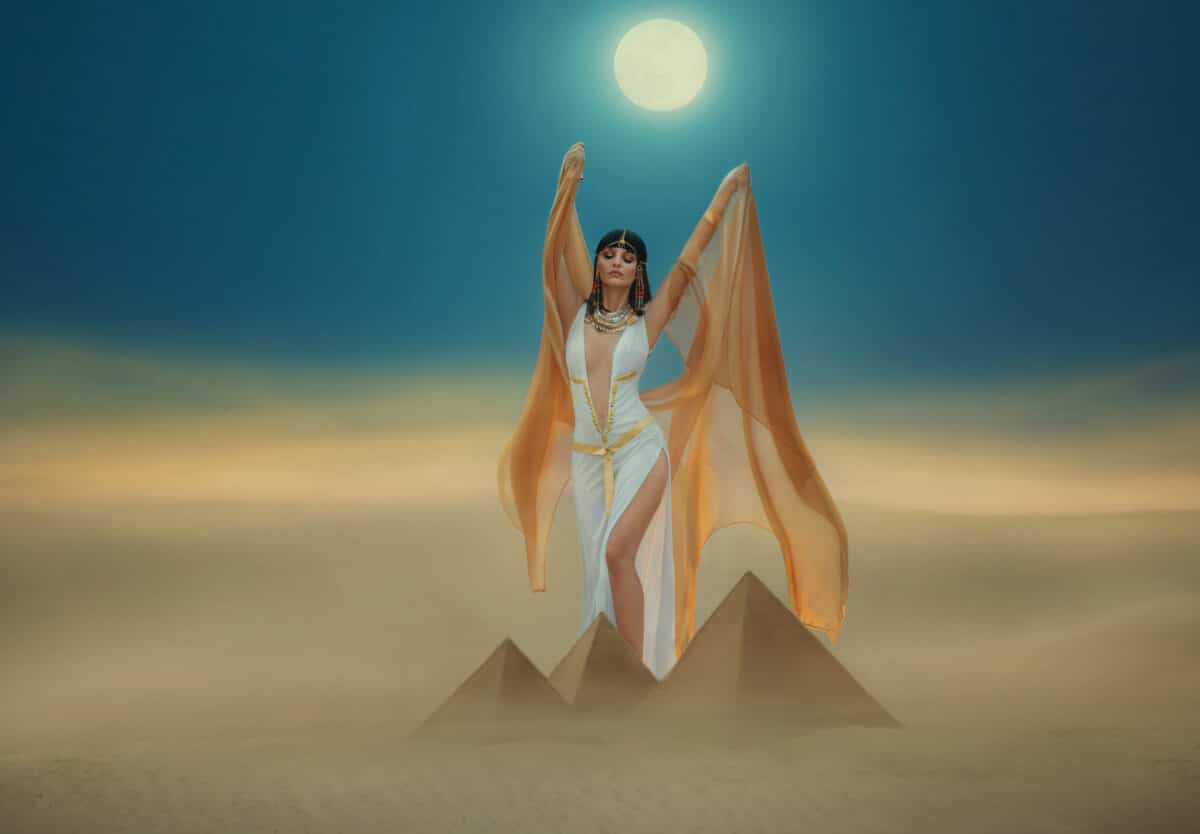 Artwork Fantasy egyptian beauty goddess Cleopatra raises hands to blue night sky, backdrop pyramid yellow sand dune, bright moon light. Orange golden cape white sexy dress. Black hair queen Nefertiti