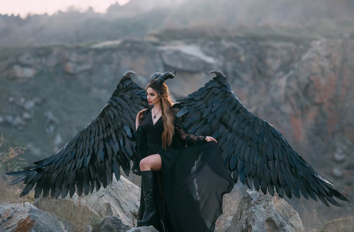 Fantasy dark magician in a black dress with huge wings and horns. Gothic model Transformation of mythical goddess Gamayun syrin. Fabolous mountains and cliffs. image outfit for Halloween party