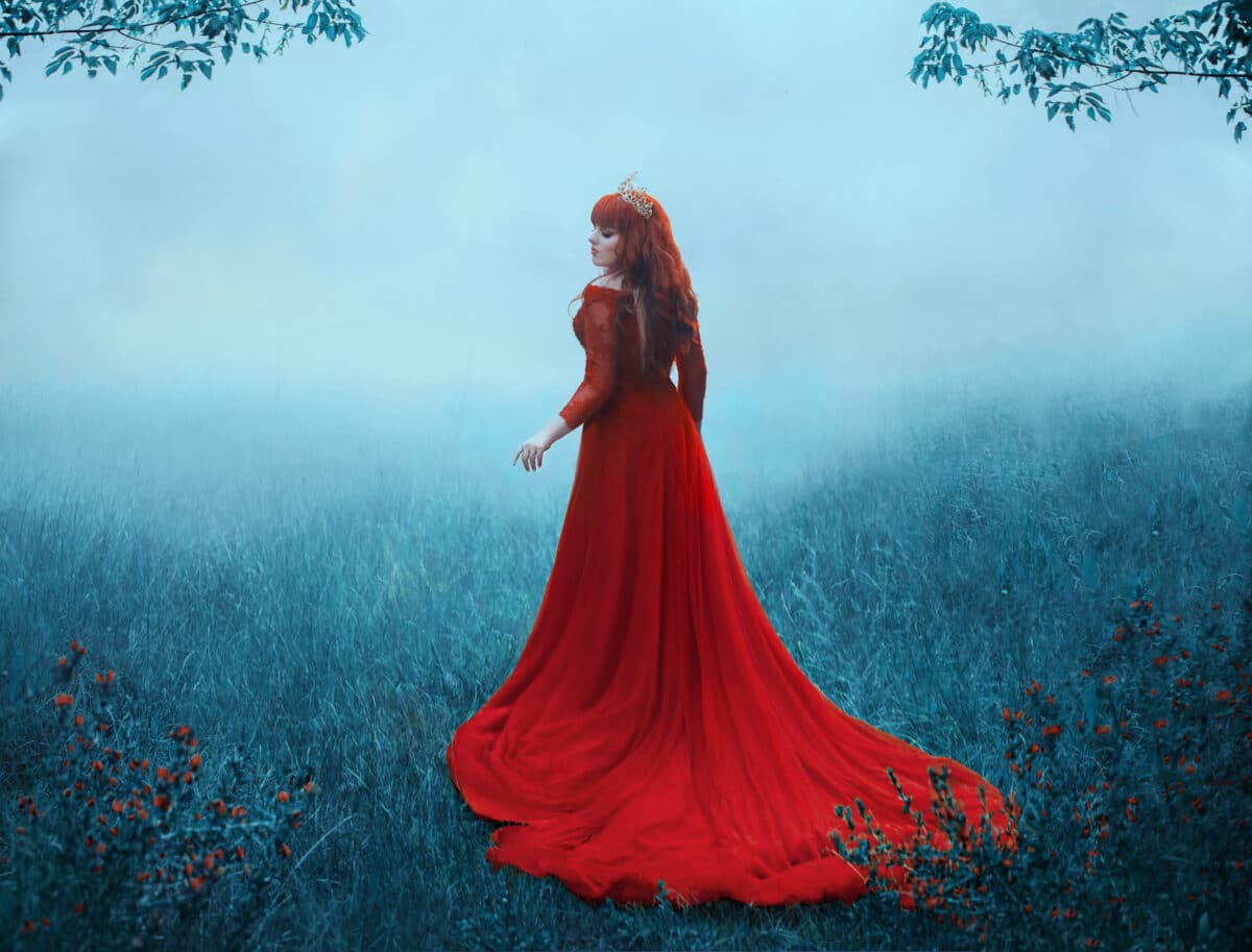The Queen in a luxurious, expensive, red dress, walks in a thick fog with a long train. A young-haired girl in a golden crown examines her possessions. The background is cold, misty autumn. Art photo.