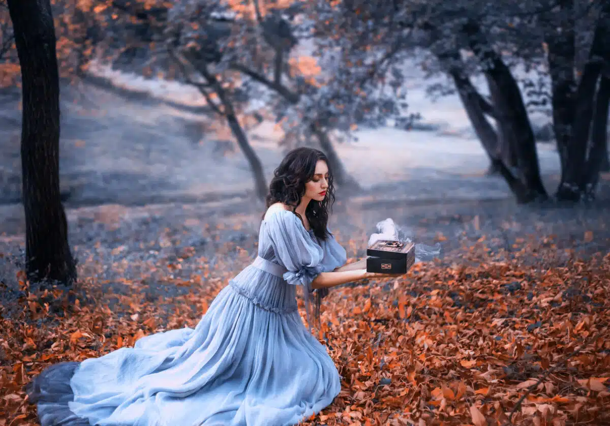 attractive brunette girl sits in a dark forest on fallen autumn orazhevyh leaves, dressed in a gray vintage dress with bare shoulders, holding in her hands an open Pandora's box full of evil, misery