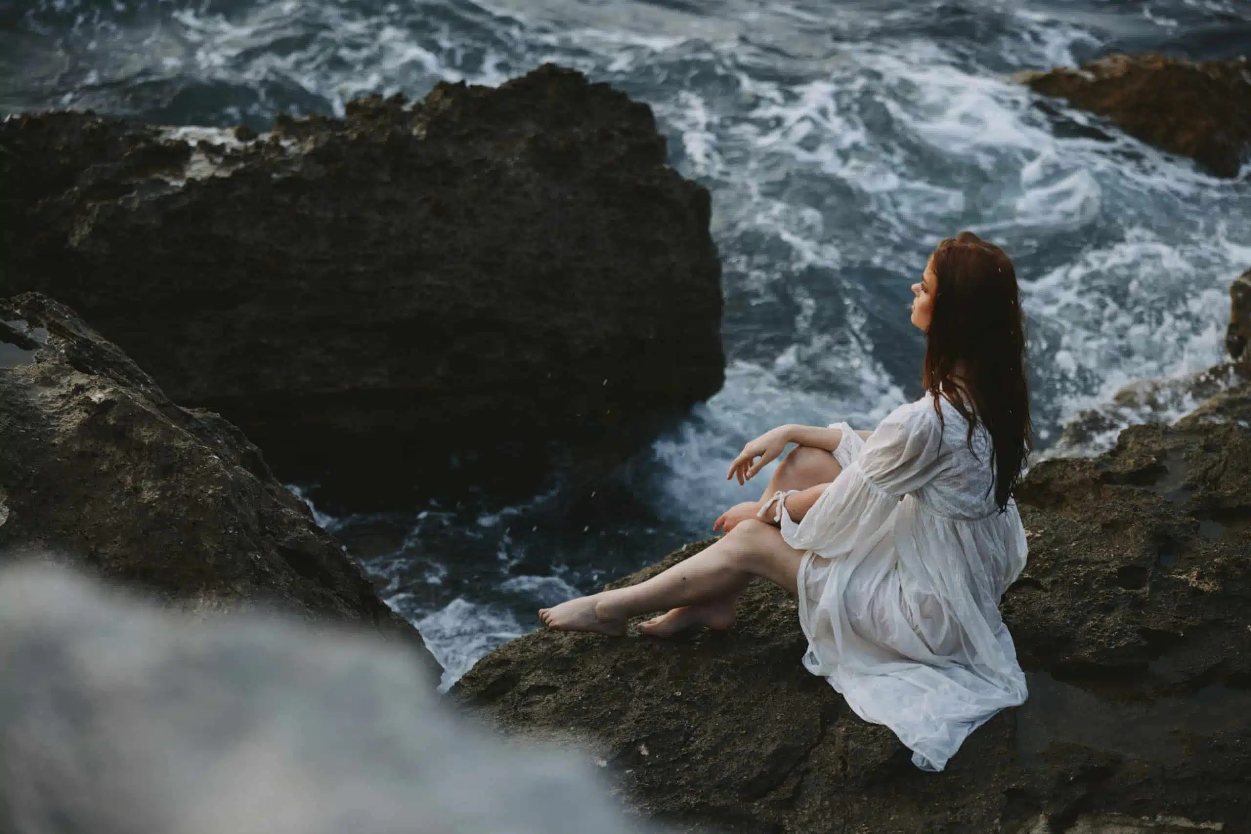 Woman in a white dress sits on a cliff barefoot.