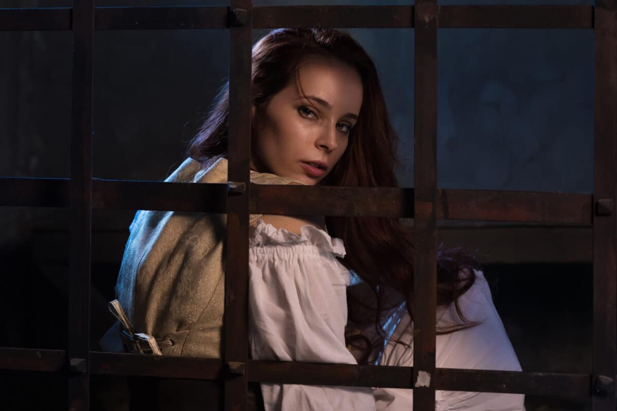 Large portrait of the odiny sad girl of the pirate in the prison cell behind an iron lattice. Dark background