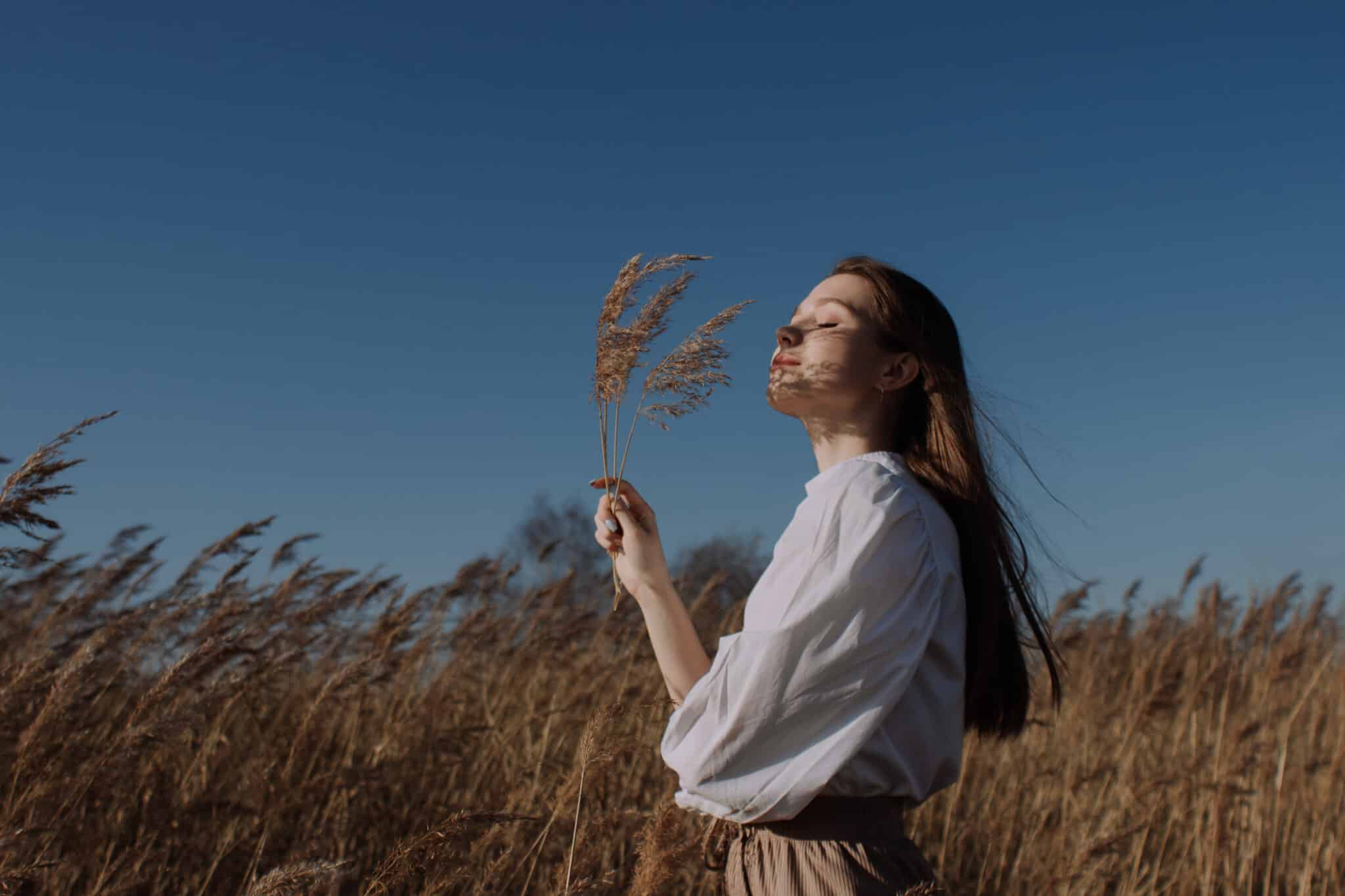 young woman with eyes closed standing in the field holding branches of dry pampas grass under the blue sky
