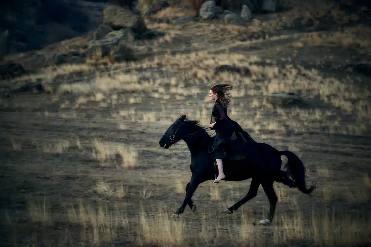 Woman in black dress riding a black horse out in the field.