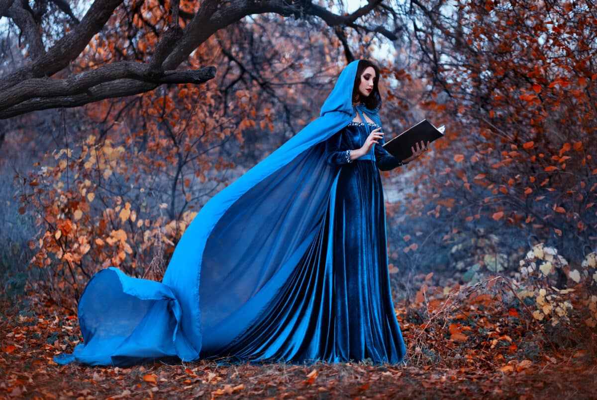 Fantasy Woman witch holds magic book in hands reads spell. Blue vintage clothes silk cape dress flies in wind. Mystery girl elf in hood. Nature autumn forest trees orange leaves. Holiday halloween art