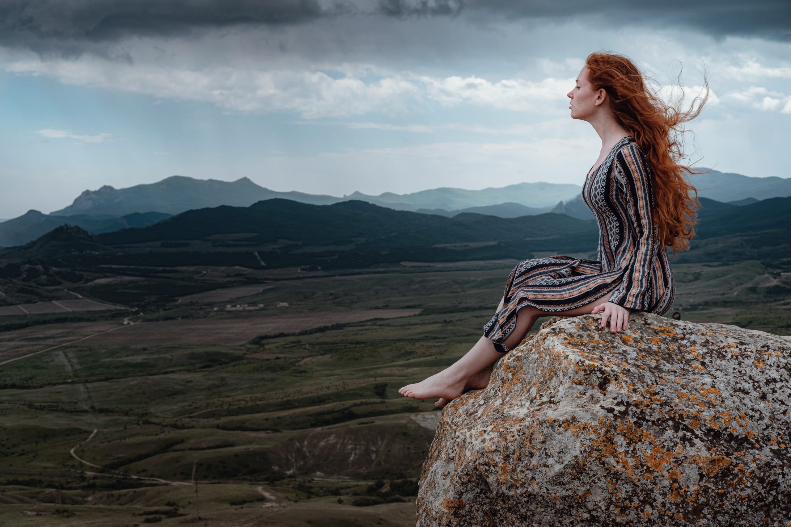 Ginger-haired woman sits on a cliff and looks far ahead.