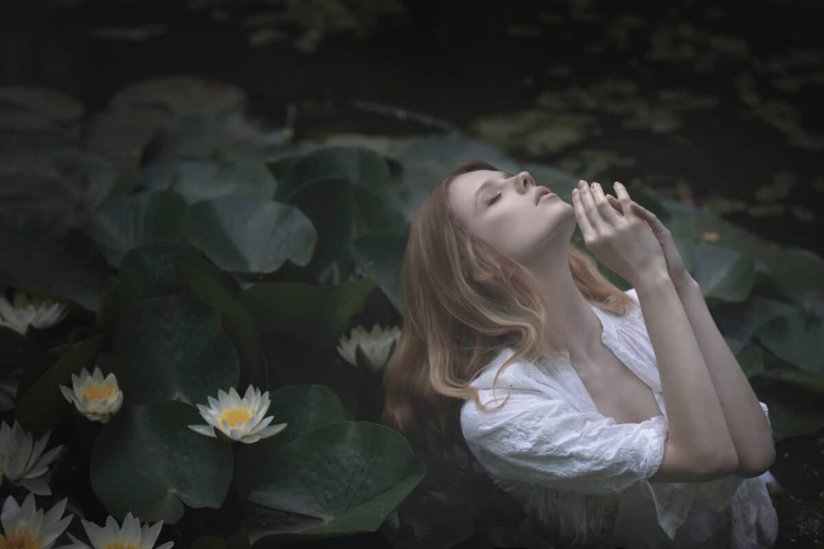 Young blonde nymph praying head raised above in dark water with white lilies