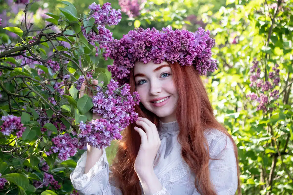 Young Girl With Long Red Hair With A Wreath Of Lilacs On Her Head In The Garden