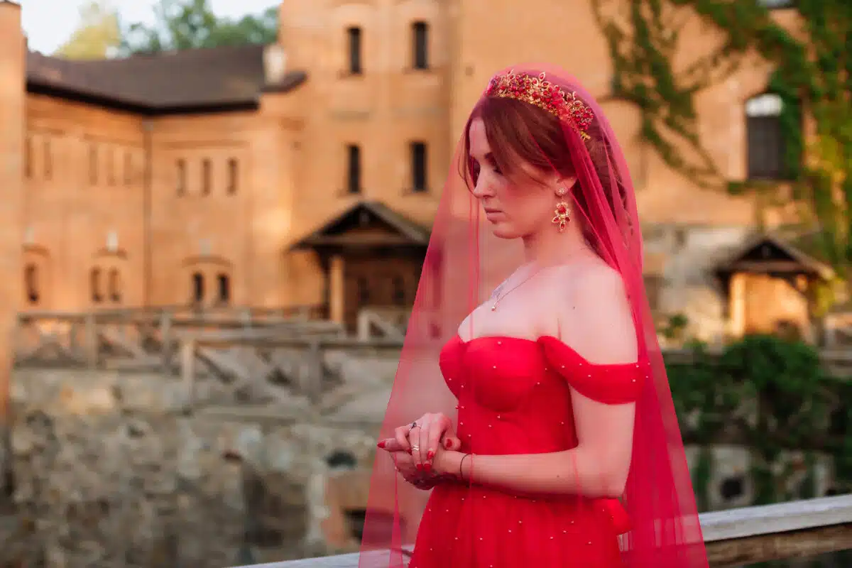 attractive but sad redhead woman in red dress wearing a diadem and a red veil outside the castle

