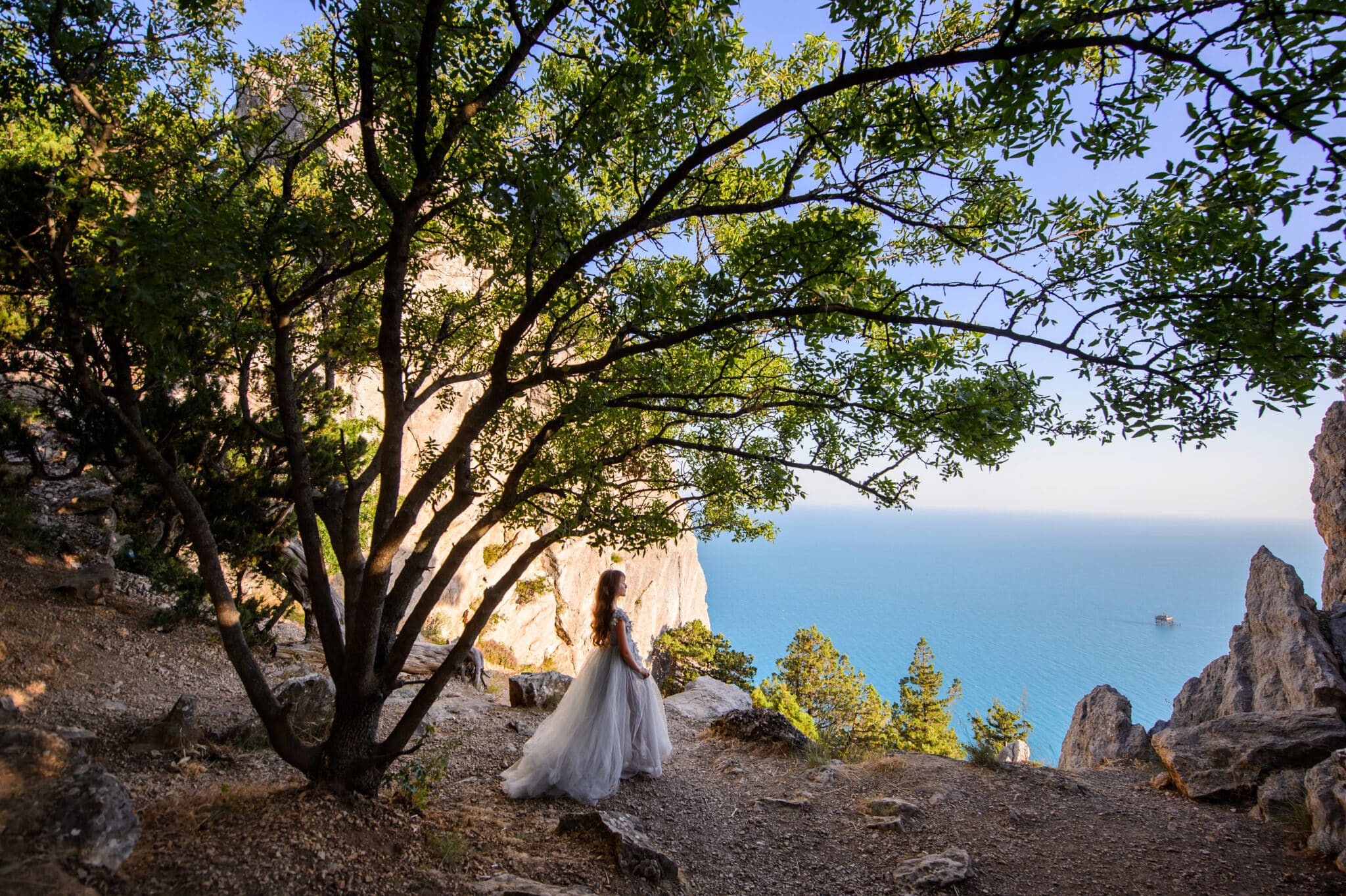 Beautiful young lady in white dress on a cliff with trees and view of the calm ocean