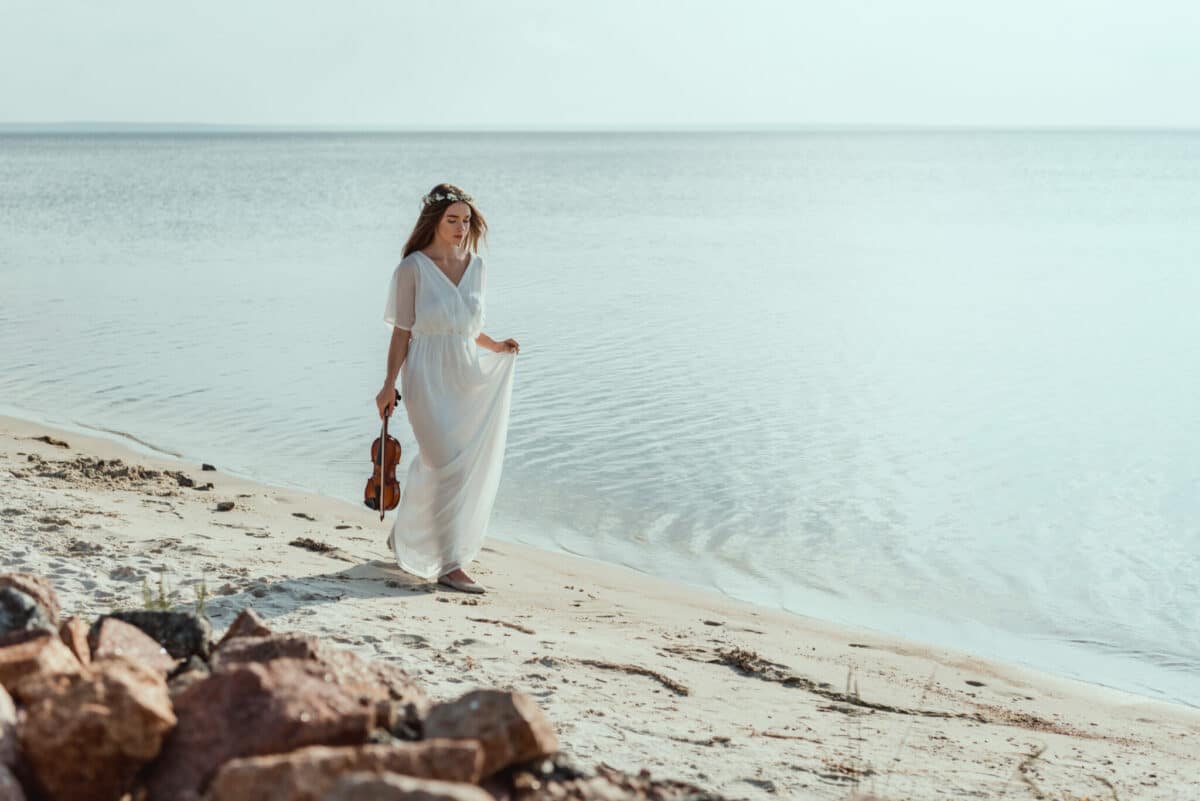 attractive young woman in elegant dress with violin walking on the beach