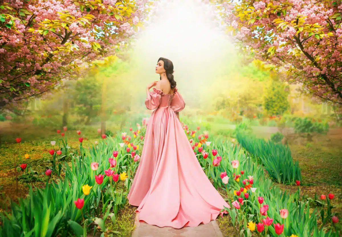 Fantasy girl princess walks in blooming spring garden flowers tulips sakura tree green grass old alley. Woman queen in long luxurious royal pink dress with train puffed sleeves vintage style art photo