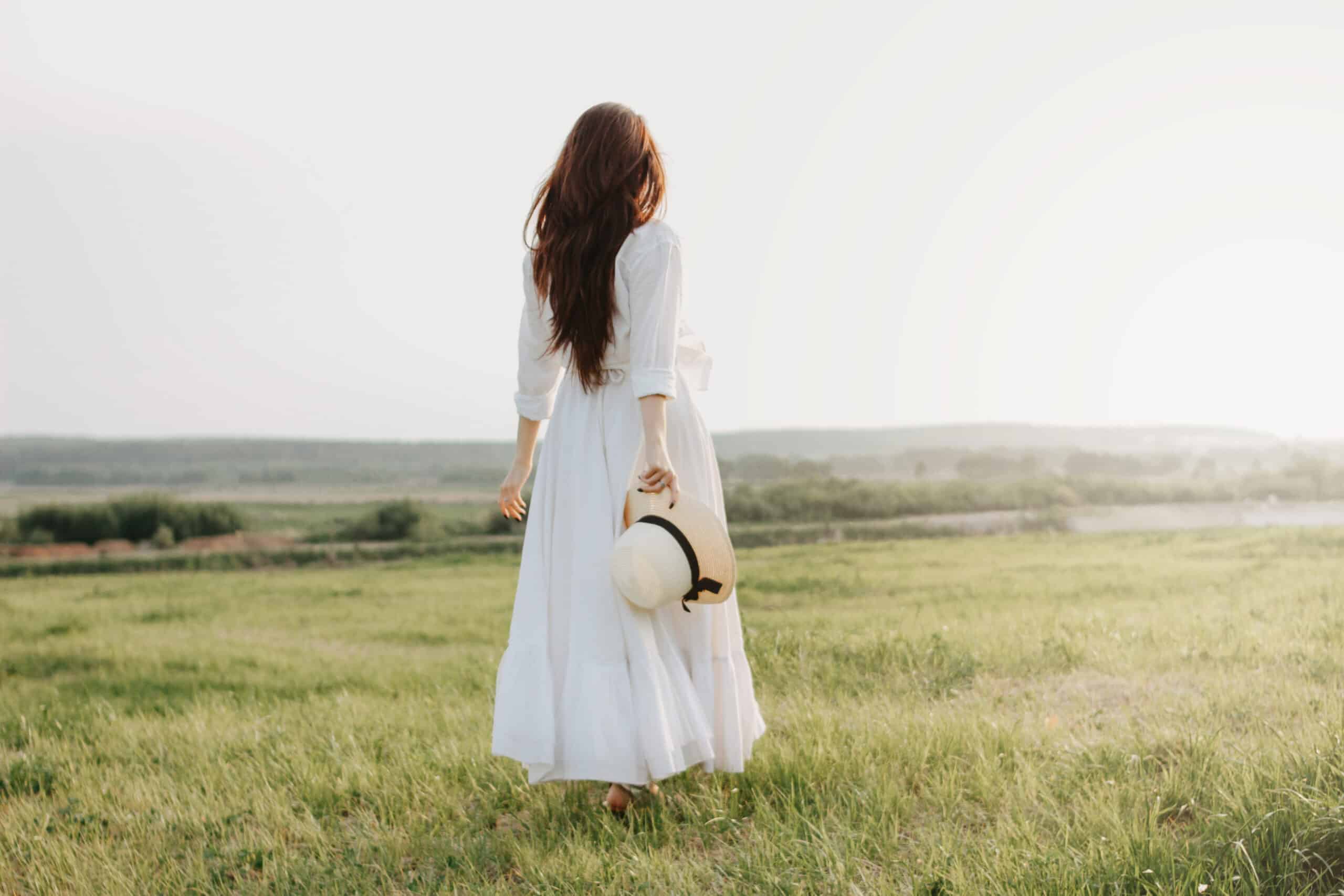Beautiful carefree long haired woman in white dress and straw hat enjoys life in nature field looking towards the sunset.