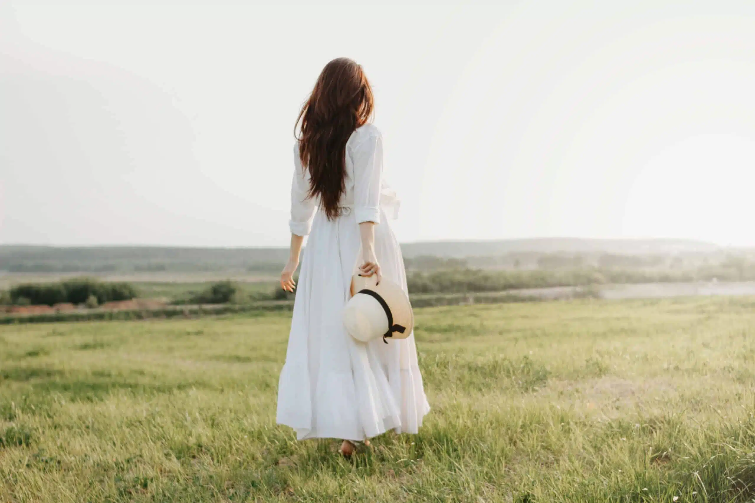 Beautiful carefree long haired woman in white dress and straw hat enjoys life in nature field looking towards the sunset.